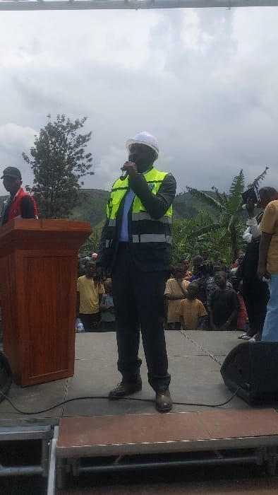 The image shows a man speaking into a microphone on a stage in Sake, Democratic Republic of Congo. The man is wearing a white hard hat and a bright yellow safety vest over a black suit. He is standing behind a wooden podium with an upright microphone. There are many people in the audience in front of him. The man on stage is speaking to the crowd. The audience is seated on the ground. There are two speakers on the ground at the right of the image. The image was taken during a cloudy day. The background shows a green grassy area with large trees, and hills in the distance.