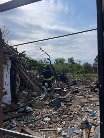 The image shows the aftermath of a fire or explosion at a property in Bilopillia, Ukraine. The location is near the intersection of Pershotravneva Street and Velyka Pereshchepinska Street. A single, unidentified firefighter, wearing a black uniform, is visible, standing in the debris, holding a fire hose. The scene is littered with the remnants of a building, including charred wood, brick, and debris. The ground is covered with dirt and dust. The sky is overcast with clouds, and there are trees and other vegetation in the background. The scene suggests destruction and recent damage, and the firefighter is possibly inspecting the damage or securing the site.