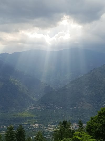 The image shows a valley in Sarsai, India, with mountains covered in lush green trees. The sky is dramatic, with dark clouds partially obscuring the sun, creating visible sunbeams that shine down on the landscape. In the valley, there are scattered houses and buildings, indicating a populated area. The foreground features the tops of evergreen trees. The overall impression is one of a majestic natural landscape with human habitation nestled within.