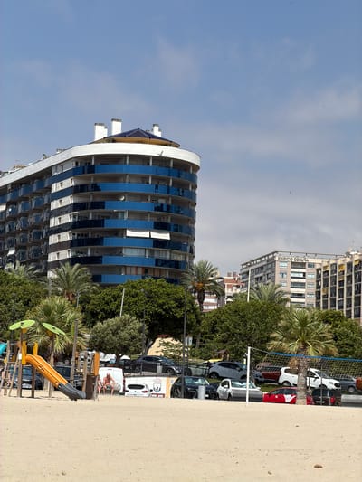 This media file shows an outdoor scene with a beach in the foreground, a playground, trees, buildings, and parked cars in the background. The image is oriented vertically, with the sky at the top and the beach at the bottom.
In the foreground, there is a sandy beach with a children's playground featuring a yellow slide and green leaf-shaped decorations. Beyond the playground, there are several trees, including palm trees.
In the middle ground, there are several parked cars, including white vans and other vehicles. A white van has a drawing on its side. Two people are sitting on a low wall near the vans. A volleyball net is visible.
In the background, there are modern buildings. The most prominent building is a curved structure with many floors and reflective blue windows. Further in the background, there is a taller, older building with many windows, some of which appear empty or damaged. The text "EDIFICIO CORUÑA" is visible on this building.
The sky is blue with some clouds. The lighting suggests it is daytime, likely sunny. The overall setting appears to be a coastal urban area, possibly a resort, in Spain, as indicated by the location context. The scene conveys a sense of leisure and urban life.