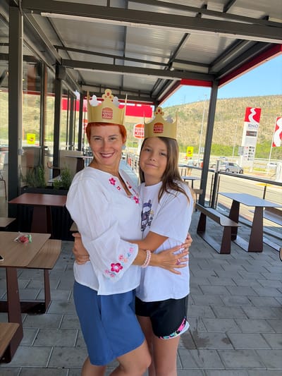 The image shows two females, an adult and a child, embracing and wearing Burger King crowns outside. They are standing on a paved area in front of an outdoor seating section of a Burger King restaurant. In the background, there are mountains, a road with cars, and a gas station with signage for "EKO". The outdoor seating area includes tables and benches. The adult has short red hair and is wearing a white embroidered shirt and blue shorts. The child has long brown hair and is wearing a white t-shirt and black shorts. The scene appears to be in Gornje Mrke, Montenegro, given the context provided.