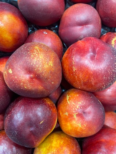 This image is a close-up, overhead shot of a pile of ripe nectarines at a market stall in Bel Air, Spain. The nectarines are primarily deep red with streaks of orange and yellow, and some are speckled with small white dots. The fruits are tightly packed together, suggesting they are for sale. The background is dark and indistinct, providing no further environmental details.