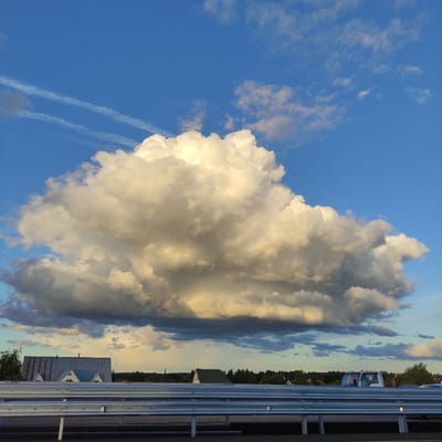The image captures a dramatic sky scene over Марусино, Russia, dominated by a massive, illuminated cumulus cloud that stands out against the vibrant blue sky. Multiple contrails streak across the upper portion of the sky, suggesting recent aircraft activity. In the middle ground, a row of houses with distinctive metal roofs and gabled windows is visible. In the foreground, a modern metal guardrail runs horizontally across the frame, indicating the viewpoint is likely from a road or elevated pathway. The overall impression is of a vast, dynamic sky juxtaposed with a rural or semi-rural landscape.