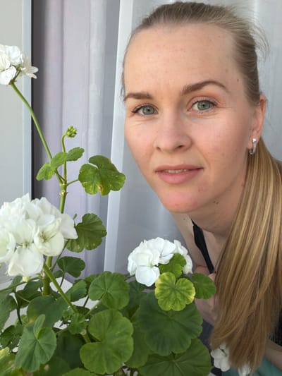 The image shows a medium shot of a woman with blonde hair and green eyes, looking towards the camera. She is partially obscured by a potted plant with white flowers and green leaves. The woman is wearing a black top and silver hoop earrings. She appears to be indoors, with a light-colored curtain in the background. The image is well-lit and in focus. There is no visible indication of the location being in the city of Кульдур, Russia.