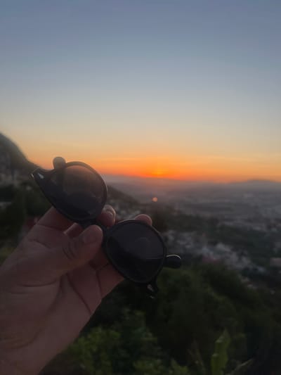 The image captures a serene sunset scene over a city, likely Tétouan, Morocco, viewed from a high elevation. The expansive sky dominates the upper portion, showcasing a dramatic gradient of colors. On the left, the sky is a soft, pale blue-grey, gradually transitioning to warm oranges and yellows towards the right, where the setting sun casts a vivid glow just above the distant horizon.

Below the horizon, the sprawling urban landscape of Tétouan is dimly visible, dotted with faint lights against the fading daylight. Hills and dark green foliage frame the lower and far-right sections of the scene, suggesting a mountainous or elevated viewpoint.

In the immediate foreground, towards the bottom right, a person's hand firmly holds a pair of black sunglasses with rounded frames. The dark lenses reflect some of the ambient light and colors. The scene evokes a tranquil moment, capturing the beauty of dusk over a city from an elevated perspective. There is no visible text in the image.