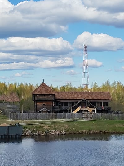 The image depicts a large wooden structure, likely a historical building or a museum, located near the water's edge. The building has a brown shingled roof, multiple windows, and a wooden fence surrounding it. In the background, there's a tall telecommunications tower and a forest of green trees under a blue sky with puffy white clouds. A body of water, likely a lake or river, is in the foreground. The image was taken in the city of Шиндалово, Russia.