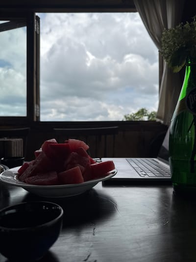 A still life photograph taken indoors at an establishment in Thôn 4, Vietnam, depicts a slice of watermelon on a white plate, a laptop, and a green bottle of Perrier water on a dark wooden table. The scene is framed by an open window overlooking a cloudy sky and lush green trees. In the foreground, a dark bowl sits on the table. The lighting is dim, suggesting it may be late in the day or the establishment is naturally dark. The window provides natural light, casting soft shadows across the scene. The overall impression is one of a tranquil, perhaps work-related, moment enjoyed with a refreshing snack.