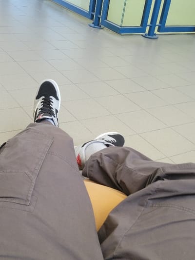 The image shows a person sitting on a floor in a public waiting area in Calais, France. The person is wearing grey cargo pants, and black and white sneakers. They are sitting on a tan colored cushion, resting their feet on a floor made of grey tiles. The person is wearing socks, and their other foot is out of view. There is a blue railing in the background with a glass partition. The floor is covered in grey tiles, and the image is taken from the perspective of the person's right leg. The image is taken at the coordinates 50.953 latitude and 1.879 longitude.