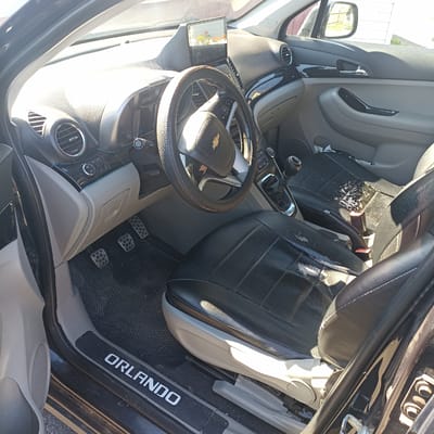 The image shows the interior of a car, specifically the driver's side, from a perspective just inside the open door. The car appears to be parked on a gravel surface, with a white building visible in the background through the side mirror. The interior is predominantly grey and black, with a black steering wheel with a Chevrolet logo, a gear shifter, and two pedals visible on the floor. The driver's seat is black with visible wear and tear on the upholstery. The dashboard features air vents, a radio or infotainment screen displaying an image of flowers, and various buttons. The "Orlando" text is visible on the door sill plate. The lighting suggests it is daytime, with strong sunlight entering the car from the right, creating bright highlights and shadows. There are no people visible in the image. The overall condition of the car's interior suggests it has been used and may be in need of cleaning or repair.