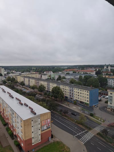 The image depicts an overcast day in the city of Piła, Poland, showing a view of a residential area and surrounding buildings. In the foreground, a long, multi-story apartment building with a light-colored roof runs diagonally across the frame. The building has a facade of yellow and red sections with numerous windows. Adjacent to this building is a street with cars, parked vehicles, a pedestrian crossing, and trees lining the side.
Further into the distance, several other apartment buildings are visible, some with yellow facades and others with a mix of colors including green and blue. Beyond the residential blocks, a denser area of buildings suggests a more commercial or older part of the city. Distinctive structures include a church with twin steeples and a large, red-roofed building. The background is filled with a dense line of trees, indicating a forest or large park bordering the city. The sky is uniformly grey and cloudy, typical of an overcast day. A few people are visible on the sidewalks and streets, walking or standing. The overall scene is one of urban residential development with natural elements present in the background.