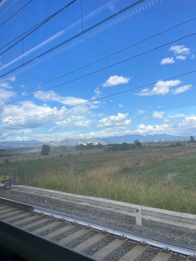The image depicts an exterior view from a train window, showcasing a rural landscape under a clear blue sky with scattered cumulus clouds. In the foreground, two parallel train tracks, consisting of steel rails mounted on concrete sleepers and ballast, extend towards the horizon. Adjacent to the tracks on the right, a low, light-colored concrete barrier runs parallel to the railway line. The immediate ground cover alongside the tracks and barrier includes a mixture of green and dry brown vegetation.

The midground presents expansive agricultural fields, characterized by varying shades of green and brown vegetation, indicative of cultivated land. Faint fencing structures delineate some field boundaries. Scattered trees are present throughout the midground, notably an isolated dark green tree towards the left-center, behind which a distinct plume of light-colored dust or smoke rises vertically from the ground. Further in the midground, distant, indistinct buildings or structures are visible on the right.

The background features a range of hazy, blue-grey mountains stretching across the horizon. Reflections of interior lighting, appearing as small white dots, are visible on the upper portion of the train window glass. The scene is located in or near the city of Afragola, Italy. No individuals or active interactions are observed.