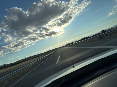 An image taken from inside a vehicle shows a view of a highway in Krivi Dol, North Macedonia. The road stretches ahead, disappearing into the distance, with a clear blue sky above. Large, dramatic clouds, illuminated by the sun, dominate the upper portion of the frame. The sun is partially obscured by a cloud, creating a bright glare with visible light rays. On either side of the highway, there are fields and rolling hills, with some trees visible in the distance. There are guardrails along the side of the road, and power lines can be seen in the landscape. The interior of the vehicle, including the dashboard and windshield, is partially visible in the foreground.