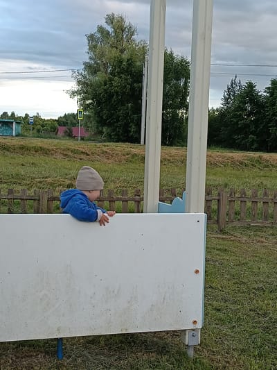 A young child wearing a grey knitted hat and a blue hooded jacket is peeking over a white structure in an outdoor setting. The child is facing to the right, with their hands resting on the edge of the structure. The scene appears to be a rural or semi-rural area, possibly Smolino, Russia. In the background, there is a wooden fence, dry grass, green trees, and a cloudy sky. There are also some structures visible in the distance, including a small building with a green facade and a bus stop sign. A yellow pedestrian crossing sign is also visible. The time of day appears to be late afternoon or early evening, with diffused light suggesting overcast weather. The child's expression is not clearly visible, but their posture suggests curiosity or engagement with something out of frame. The white structure the child is leaning on looks like it could be part of a playground or an outdoor seating area.