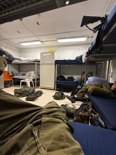 The image shows the interior of a military barracks room, likely located at the Kiryat Gat base, Mazkeret Batya, Israel, as per the provided coordinates. The room is sparsely furnished, with two sets of bunk beds lining the walls. A white refrigerator stands against the wall opposite the beds. A lone person is lying on the top bunk, wearing olive green military fatigues. The person's face is obscured. The room is cluttered with military gear and clothing strewn about the floor. Other military personnel may be present, but only a lower body and feet of one person are visible in the foreground. The room has a white ceiling with exposed wires and pipes. A single fluorescent light fixture is visible in the ceiling. The walls are painted white, and the floor is covered in a light-colored tile. The room is dark, indicating a lack of windows. A red fire alarm light is visible near the ceiling. The image is taken from a low angle, possibly from the bottom bunk. A small, orange chair is visible in the background, to the left of the refrigerator. The image captures the scene from a first-person perspective, as if the viewer is also lying on a bed. A small, round, brown object, possibly a helmet, is visible in the bottom right corner of the image. The photograph was taken indoors, at night, in an enclosed military barracks setting.