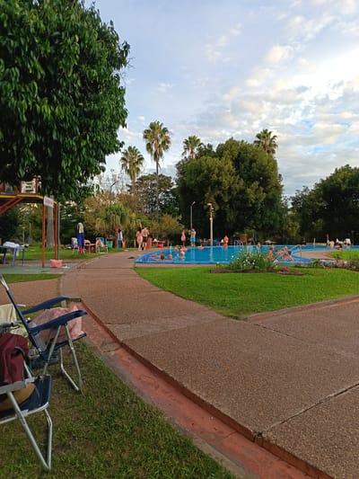 The image depicts a public swimming pool in Termas del Daymán, Uruguay. The pool is filled with people, primarily adults, swimming and relaxing. Surrounding the pool is a paved area with grass patches, flowers, and trees, including palm trees. Several lounge chairs are scattered around the pool area. A two-story lifeguard tower is visible in the background. The sky is partly cloudy. In the foreground, two unoccupied folding chairs are placed on the grass.