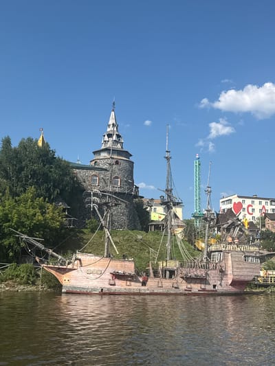 This image captures a scene in Satka, Russia, featuring a large, elaborate pirate ship replica docked on a body of water. The ship, predominantly constructed of wood with a reddish-pink hull and detailed rigging, occupies the foreground. Behind the ship, a stone tower with a conical roof, reminiscent of a castle or fortress, stands on a grassy hill. Further in the background, a tall, green amusement park ride, possibly a drop tower, is visible against a clear blue sky with scattered white clouds. Several other buildings, some with colorful facades and architectural elements, are also present on the hillside. The overall environment appears to be a recreational area or theme park, with the pirate ship serving as a central attraction.
