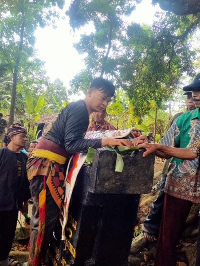 An outdoor scene in Menjing, Indonesia, features at least five men gathered around a dark, unpolished stone structure. In the foreground, a man with short, dark hair wears a dark long-sleeved shirt, a yellow waist sash, and a patterned sarong-like garment with black, orange, and red geometric designs and red and yellow tassels. A white fabric with red and black patterns and tassels is draped over his left shoulder. He leans forward, manipulating green banana leaves placed on the flat top surface of the stone structure. The structure consists of a darker base and a lighter, rough-hewn top block. Partial text, including "NG" and "MENJING," is visible on the lower section of the base. To the right, a second man wearing a green patterned shirt and a dark blue cap has his right hand positioned near the stone structure. To the left, a third man wearing a dark shirt and a patterned headband observes the activity. Additional figures are visible in the background among the lush green vegetation, which includes tall trees with dense foliage and banana plants. The scene is illuminated by bright, overhead ambient light.