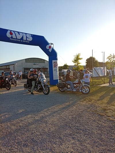 A group of motorcyclists are gathered under a large blue inflatable archway bearing the "AVIS Taverne e Arbia" logo. The archway is positioned in front of a long, low-lying building with a grey facade, possibly a warehouse or industrial facility. Several people are visible on and around motorcycles, with some individuals standing and conversing. One motorcycle, a black Harley-Davidson, is prominent in the foreground with two people on it. Another similar motorcycle is to its left. The ground is covered in gravel, and a grassy area is visible to the right, separated by a fence. The sky is clear and blue with the sun low in the sky, casting long shadows. The scene appears to be an outdoor event or gathering in Arbia, Italy, possibly related to a motorcycle rally or community event sponsored by AVIS.
