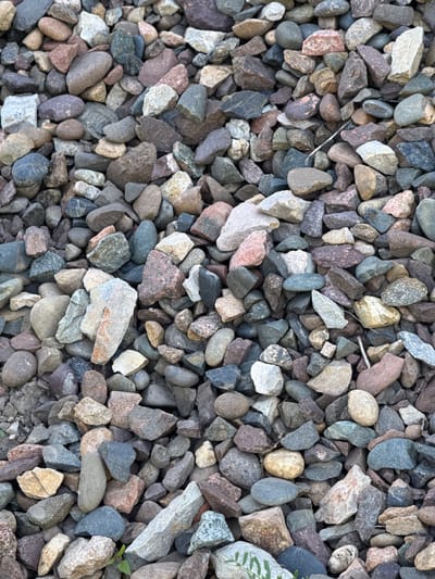 The image provides a close-up, overhead perspective of a dense aggregation of gravel, likely serving as ground cover in an outdoor environment in Зыково, Russia. The scene is static, depicting no human subjects or active events.
The gravel consists of a myriad of small to medium-sized rocks and pebbles, showcasing a rich and varied spectrum of colors. Predominant hues include various shades of gray, dark blue, muted green, earthy brown, reddish-pink, and off-white or cream. The rocks display a range of shapes, from sharply angular and fractured pieces to more rounded and smoothed stones, indicating diverse origins and degrees of erosion. Their surfaces appear rugged and textured.
Notable details within the composition include sparse evidence of plant life; small green shoots are visible emerging from between the stones, particularly towards the bottom right of the frame in the original image. In one of the cropped sections (top left crop), a thin, silvery, metallic strand or wire can be seen nestled among the pebbles. The lighting suggests daylight conditions, likely diffused or overcast, as there are no distinct, hard shadows. The rocks themselves appear dry. No discernible text is visible anywhere in the image.