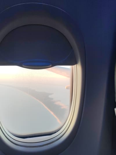 The image is a view from an airplane window, looking out at a hazy sky. The window frame, made of dark blue plastic, is visible in the foreground, with a pull-down shade obscuring the top portion of the window. The shade has a light blue handle.

Outside the window, the sky appears to be filled with a soft, diffused light, suggesting either sunrise or sunset. A faint, rainbow-like arc of color is visible in the sky, possibly a reflection or a weather phenomenon. Below the sky, a layer of clouds or landmass is vaguely discernible, appearing as dark shapes against the lighter sky. The overall impression is one of tranquil travel, with the soft lighting and the muted colors of the sky creating a peaceful atmosphere. There is no visible text in the image. The scene is likely taking place during a flight over Russia, as indicated by the location context of Khrabrovo, Russia.