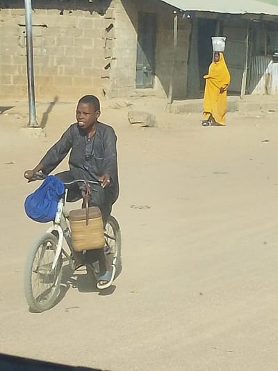 The image depicts an outdoor scene in the city of Mariri, Nigeria, on an unpaved dirt road under clear daylight. In the foreground, a young male with dark skin and short dark hair, wearing a dark grey long-sleeved tunic and light-colored sandals, rides a white bicycle from right to left. His mouth is open, suggesting speech or exertion. A blue fabric bag is tied to the bicycle's handlebars, and a tan, rectangular insulated container hangs from the bicycle frame below the handlebars.
In the mid-ground, on the opposite side of the road, a female figure walks away from the viewer, clad in a bright yellow full-body covering that obscures most features. She carries a silver-colored bucket or container balanced on her head.
The background features simple, single-story buildings constructed of light-colored stone or brick, with at least one having a corrugated metal roof. A tall, dark metal pole stands on the left side of the road. Strong shadows indicate bright sunlight. No direct interaction between the two individuals is evident.