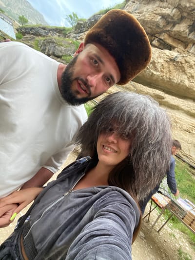 A man and a woman are taking a selfie outdoors. The man is wearing a white t-shirt and a brown fur hat. He has a beard and is looking at the camera with his mouth slightly open. The woman is wearing a gray velour tracksuit and her hair is covering her face. She is smiling at the camera. They are holding hands. In the background, there are rocky cliffs and some greenery. There is also a table with items on it and another person in the background. The location is identified as the city of Salta, Russia.