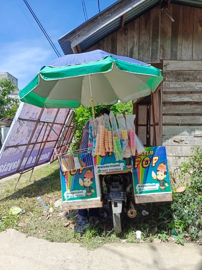 An outdoor scene in Meuliek, Indonesia, features a mobile ice cream cart parked on a grassy area beside a weathered wooden building. The cart, adorned with colorful graphics of an ice cream cone and a child, displays the words "ES KRIM POP" and a phone number. It is covered by a large green and blue umbrella. Several bags of colorful ice cream cones are neatly arranged on the cart. A white wire basket hangs from the side of the cart, holding paper cups. Behind the cart, a motorcycle serves as the base, with its exhaust pipe visible. A large banner, leaning against a metal frame, displays images of people and text in an Indonesian language. The ground around the cart is littered with plastic bottles and debris. Lush green vegetation grows around the building and the cart. The sky above is a clear blue, with power lines crisscrossing overhead.