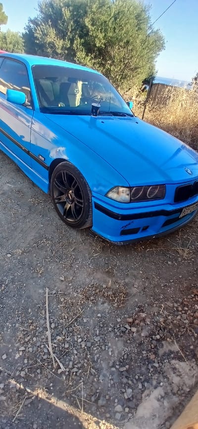 A bright blue BMW E36 coupe is parked on a gravelly surface outdoors. The car is angled towards the viewer's right, with its front end prominently featured. The vehicle has aftermarket black rims and a custom front bumper with a sticker on the lower left side. A cup with a straw sits on the dashboard. In the background, dry grass and vegetation are visible, along with a hint of a tree line and a clear blue sky. The overall impression is that of a car parked in a rural or undeveloped area, possibly in the vicinity of Chersonisos, Greece, given the characteristic dry Mediterranean vegetation.