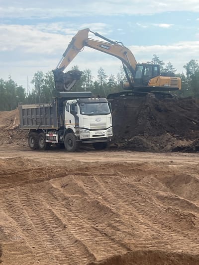 A yellow SANY SY330C excavator is positioned on a large pile of dark soil, with its boom extended and bucket poised over the bed of a white FAW Trucks dump truck. The dump truck, which has "FAW TRUCKS" written on its side, is parked adjacent to the soil pile and is facing to the right of the frame. Its bed is empty. The scene appears to be a construction or earthmoving site, likely in Shapki, Russia, given the presence of birch trees and the overall landscape. The ground in the foreground is composed of sand with visible tire tracks from heavy machinery. The sky is partly cloudy, with patches of blue visible between white clouds. There is a single person visible inside the excavator's cab.