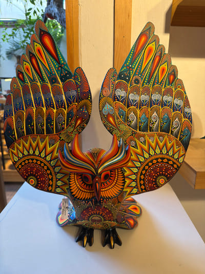 This media file features a vibrantly painted wooden sculpture of an owl, characteristic of Zapotec alebrije folk art from San Martín Tilcajete, Mexico. The owl is depicted with large, outstretched wings, adorned with intricate, psychedelic patterns and a rich palette of colors including brilliant oranges, yellows, greens, blues, reds, and black, highlighted with gold-like accents.
Every section of the owl, from its feathers to its body, is covered in meticulously painted motifs such as concentric circles, spirals, teardrop shapes, dots, and geometric patterns, creating a highly detailed and captivating visual. The owl's stylized face features prominent eyes and a sharp beak, while its black, clawed feet are visible on the right side of the sculpture.
The sculpture is displayed indoors, mounted against a light-colored wall. Wooden beams or shelves are visible in the background, and green foliage can be glimpsed through a window or opening in the lower left, suggesting natural light.
A small tag is attached near the owl's feet, displaying the following text: "BUHO DE 4 PIEZAS" (Owl of 4 pieces), "VIVE", and "PRECIO $5,200.00". The intricate craftsmanship and vibrant aesthetic are hallmarks of Oaxacan artisanal traditions.