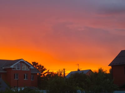 This is a horizontal, eye-level, full shot of a sunset over the town of Фронтовики Снт, Russia. The sky is a vibrant gradient of orange and pink, with a few scattered clouds. In the foreground, there are several houses with red brick exteriors and dark roofs, surrounded by lush green trees. A power line with a pole and antenna is visible in the middle ground. The overall scene is peaceful and serene, capturing the beauty of a rural sunset.