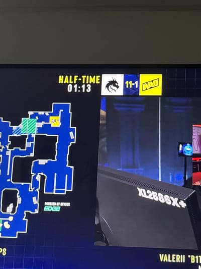 This media file appears to be a screenshot or recording of a digital display, likely from a computer monitor or a screen showing a video game interface. The primary subject is a game's user interface, displaying elements commonly found in competitive esports, specifically a tactical shooter like Counter-Strike: Global Offensive.

The display shows a score of "11-1" next to a logo that reads "NAVI," indicating this team is leading significantly. Below this, there's an icon of a dragon and the text "HALF-TIME 01:13," suggesting the game is at a midpoint with a small amount of time remaining. A minimap of a game level is prominently featured, colored in blue, with specific areas marked with green and blue striped patterns, and a yellow area labeled "A." Text at the bottom right reads "POWERED BY SKYBOX EDGE."

To the right of the game interface, parts of what appear to be hardware are visible. A black object with the text "XL2586X+" is seen, likely a model number for a monitor or computer component. Above this, yellow text reads "VALERII 'B1T'," which is a player's name or handle. The background is dark, with some blurred elements suggesting a room or studio setting, possibly where the game is being played or streamed. The overall impression is a snapshot of a professional esports match in progress, with the score indicating a dominant performance by the NAVI team. The context of Gothenburg, Sweden, is noted, but there are no visual cues within the image itself to confirm this specific location.