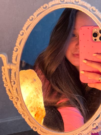 This media file is a selfie taken in a mirror. The subject is a person, reflected in an oval-shaped mirror with an ornate, white, carved frame. The person is holding a pink phone with a polka dot pattern and a raised camera module. The phone is positioned in front of their face, obscuring much of it, but their mouth and one eye are visible. Their fingernails are painted with a light color and white polka dots. The person is wearing a pink top.
Reflected in the mirror, behind the person's head, is a luminous object that appears to be a Himalayan salt lamp, emitting a warm, orange-yellow glow. The background of the reflection is a blurred blue surface, possibly a wall or fabric. The overall lighting suggests an indoor setting, possibly in the evening or in a dimly lit room, given the prominence of the salt lamp's glow. The mood conveyed is casual and personal, typical of a mirror selfie. No text is visible in the image. The location context provided is Prikubanskiy, Russia.