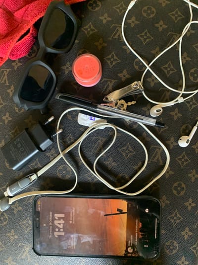 The image displays an overhead view of various personal items arranged on a brown surface embossed with the Louis Vuitton monogram pattern. The scene is located in Kubi A, Nigeria. In the upper left, a portion of a red microfiber cloth or towel is visible next to a folded pair of black sunglasses. Below the sunglasses, a small clear plastic jar containing an orange, viscous substance (likely lip balm or gloss) is present. To the right of these, a black ballpoint pen lies horizontally, with a small white rectangular piece of paper beneath it, displaying faint blue text. A silver nail clipper and two brass-colored keys on a silver keyring are situated above the pen. A black phone charger adapter with two electrical prongs is located on the mid-left. Two sets of white wired earphones are scattered across the surface; one set is loosely coiled near the top right, while the other is connected to the bottom edge of a black smartphone. The smartphone, positioned horizontally at the bottom of the frame, has a visibly cracked screen and is illuminated, displaying "Thursday, 9 October", "1:41", "Location Access Needed", and multiple notification icons. The screen's background image depicts a sunset or sunrise with a silhouette of vertical structures.