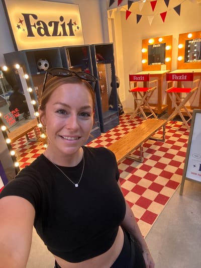 A woman with blonde hair and sunglasses perched on her head takes a selfie in a themed indoor area. She is wearing a black crop top and a necklace with a white pendant. The background features a "Fazit" logo on a lit sign, lockers, a soccer ball, red director's chairs with "Fazit" printed on them, and a checkered red and white rug. In the background, a person is visible in silhouette. A sign with "GET YOUR FAN FACE ON!!!" is also visible. The setting appears to be a promotional event or display, possibly related to sports, given the soccer ball and locker room aesthetic. The overall environment suggests a public indoor space.