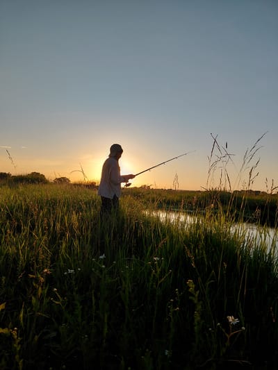 A person is silhouetted against a bright sunset while fishing in a grassy field near a body of water. The person, wearing a cap and long-sleeved shirt, stands in tall green grass holding a fishing rod, with the line extending towards the water. The sun is low on the horizon, casting a warm glow and creating long shadows. Trees and foliage are visible in the distance. The overall scene depicts a peaceful, natural setting during a sunset.