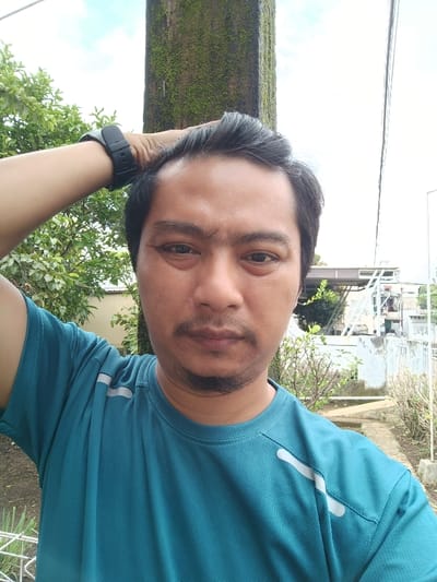 An adult male is depicted in an outdoor selfie taken in Limbangan Sari, Indonesia. The man, of apparent Asian descent, has dark hair, a mustache, and a soul patch, and is looking directly at the camera. He wears a teal short-sleeved athletic shirt featuring white diagonal stripes on the right shoulder and a black smartwatch on his left wrist. His left hand is raised, with fingers resting on the top of his head. The background on the left features a tall, vertical concrete pillar covered in green moss. Green foliage from trees and bushes occupies the mid-ground left. In the upper right, overhead power lines are visible against an overcast sky. The mid-ground right shows a white wall or fence, sparse vegetation, and a light-colored building with a dark roofline further in the distance.