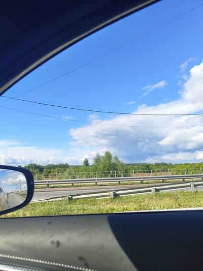 The image is a view from inside a vehicle, looking out the window and a side mirror. The scene appears to be along a highway or main road, with a guardrail separating lanes of traffic. Beyond the guardrail, there is a grassy embankment, followed by a treeline and a vast expanse of blue sky with scattered white clouds.

The side mirror reflects a road, other vehicles, and the sky. The interior of the car is visible in the foreground, including the car's door panel and a portion of the roof. The overall impression is of a sunny day, possibly in a rural or semi-rural area, given the presence of trees and open sky.

It is not possible to determine the exact location of the image within the city of Фруктовая, Russia, based solely on the visual information provided.