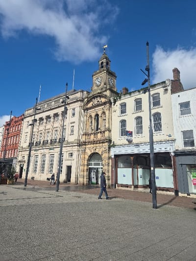 The image displays an outdoor urban scene in Hereford, United Kingdom, specifically depicting the Hereford Town Hall and adjacent buildings, likely viewed from St Owen Street or High Town, under a bright blue sky with scattered white cumulus clouds.
The central and most prominent structure is the multi-story Hereford Town Hall, constructed of light-colored stone. It features a distinguished clock tower topped with a gold weathervane. The clock face indicates a time of approximately 12:45. The building exhibits ornate architectural details including arched entrances, classical columns, statues within niches, and multiple windows. To the immediate left of the Town Hall is a section of Hereford Shirehall (now Council Offices), a similarly light-colored building with a classical facade, numerous windows, and a balustraded upper section. Further left, a multi-story building constructed of orange-red brick is partially visible.
To the right of the Town Hall, a sequence of multi-story buildings extends. The first building, directly adjoining the Town Hall, is light-colored and features several windows; an upper window displays a "TO LET" sign. Its ground floor presents a recessed, ornate storefront with large windows covered by white paper and blue signs. To the far right, another white-painted building with a grey ground-floor retail unit is partially observed.
The foreground and midground consist of a paved public area with sections of grey and reddish-brown paving stones. Several tall, black, multi-armed streetlights are interspersed throughout the scene, casting discernible shadows to the right.
Approximately six human figures are present. In the mid-foreground, a male individual wearing a dark jacket and jeans walks from left to right across the paved area. Three other indistinct figures are visible walking in the mid-left background near the Shirehall building. A single figure is partially obscured by a streetlight pole on the right side. Additionally, several pigeons are scattered on the paved ground in the mid-left. No direct interactions between individuals are evident.