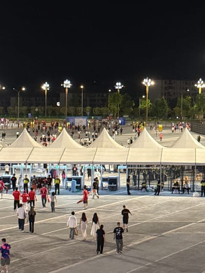 The image shows an outdoor event at night in Longxing, China. People are passing through white tents, possibly security checkpoints. The ground is marked with white lines, resembling a running track. Streetlights illuminate the area. Buildings and trees line one side of the event space.