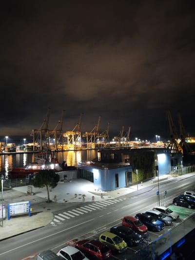 A nighttime scene shows a harbor with numerous cranes illuminated against the dark sky. Reflections of light shimmer on the water. A red tugboat is docked near the cranes. Adjacent to the water, a modern blue building is partially covered in greenery. A street runs parallel to the harbor, featuring parked cars, pedestrian crossings, and streetlights. The cloudy sky dominates the view.