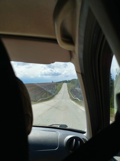 The image captures a first-person perspective from inside a vehicle, looking out through the front windshield onto a rural road in Gol'yany, Russia. The view is framed by the car's interior components: the dark dashboard and air vents at the bottom, a visible seatbelt crossing the lower right, and the car's A-pillars and sun visor framing the top and sides. Part of a dark headrest is also visible on the left.
The asphalt road stretches straight ahead, appearing to go uphill before disappearing over a crest in the distance. White guardrails line both sides of the road, though the one on the right is partially obscured by the car's interior. On the left, beyond the guardrail, lies a vast expanse of tilled brown earth, indicating agricultural fields. Further back on the left, some sparse green vegetation and trees are visible. To the right of the road, a line of trees and denser foliage can be seen, with some bare branches suggesting early spring or late autumn.
The sky above is mostly overcast with scattered white and grey clouds, suggesting daytime conditions. The overall impression is one of a journey through a peaceful, open landscape. No people are explicitly visible, and there is no discernible text within the image.