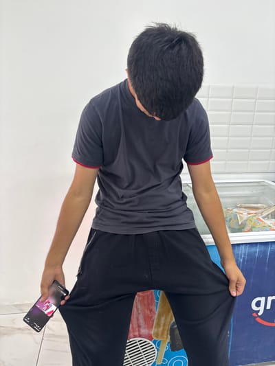 A person is angled from behind, facing right, wearing a gray t-shirt with red trim, and black pants. The left arm is extended holding the pants leg. The right arm is hanging down, holding a smartphone. The background includes a white wall, a vertical ice cream freezer with graphics, and tiled floor.