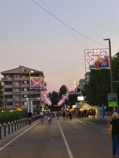 The image depicts a street scene in San Salvo, Italy, likely during an evening event, given the decorative lights. A paved street runs through the center of the frame, with sidewalks on either side. Along the street, there are several large, square-shaped light installations suspended above the road, each featuring a colorful, symmetrical design, resembling flowers or stars, illuminated with white, blue, and red lights. Streetlights and trees are also visible.
On the left side of the street, a multi-story building with a grey facade and numerous windows stands. On the right side, more buildings and trees are present, with some shops or businesses visible. A bright green sign with text and an image, possibly an advertisement, is mounted on a post on the right.
Numerous people are walking along the sidewalks and street. Most are casually dressed, suggesting leisure activity. Some are walking in groups, while others are alone. The overall atmosphere appears lively and social. The sky is a pale gradient of blue and purple, indicating dusk or early evening. The presence of decorative lights suggests a festival, celebration, or summer evening illumination.
