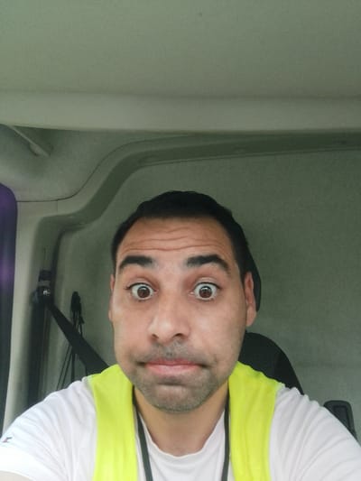 A man with dark hair and a concerned expression is inside a vehicle, possibly a truck cab, looking directly at the camera. He is wearing a white t-shirt and a bright yellow safety vest over it. The interior of the vehicle is visible behind him, with grey paneling and a seat. The lighting suggests it is daytime. The image is a selfie, taken from a close-up perspective, focusing on the man's face and upper torso. No specific details about the city of Ottersberg, Germany, are present in the visual information of the image.