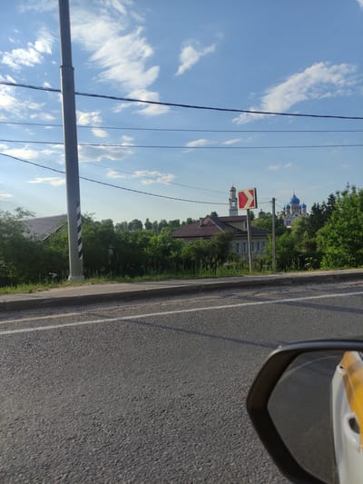 This image depicts a roadside scene in the city of Rogachovo, Russia, under a bright blue sky with scattered white clouds. In the foreground, asphalt pavement of a road is visible, with a white line marking the lane. A sidewalk runs alongside the road, bordered by grass and dense green bushes. A tall, grey utility pole with black and white diagonal markings stands prominently on the left side of the frame.
Further back, a house with a dark, sloped roof is partially obscured by the foliage. A directional sign indicating a turn to the left with a red and white chevron pattern is mounted on a pole. In the distance, a cluster of green trees forms a backdrop. Dominating the far background are two domed buildings, one with a golden dome and bell tower, and another with multiple bright blue domes, characteristic of Russian Orthodox churches. The reflection of a yellow and white object, possibly part of a vehicle, is visible in the lower right corner of the image, suggesting the photo was taken from a car.
