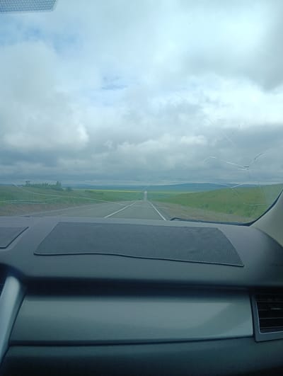 The image is a perspective view from inside a vehicle, looking through the windshield. The windshield has several cracks, most notably a large, spiderweb-like fracture across the upper right quadrant and a long, horizontal crack extending across the lower portion. Outside, a two-lane asphalt road stretches into the distance, bordered by green fields and rolling hills under a cloudy sky. The dashboard of the vehicle is visible in the foreground, with a textured speaker grille and a dark textured panel. The scene appears to be a roadside view in a rural or semi-rural area, possibly near Jerdovka, Russia, given the context. There are no people or discernible actions taking place.