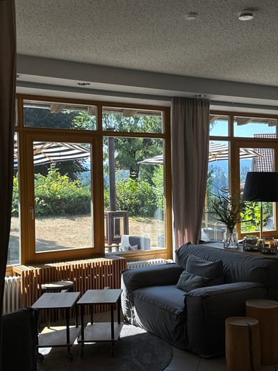 The image shows a living room interior with a large window overlooking a garden. The room features a dark grey sofa, two small wooden side tables on wheels, a wooden radiator cover, and a lamp on a shelf. Outside, the garden is lush with green trees and bushes, and two striped umbrellas are visible. The sun is shining brightly, casting shadows across the room. The location is described as St. Märgen, Germany. There are no people visible in the image.