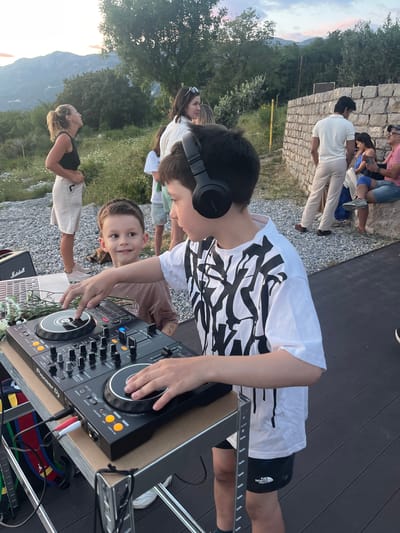 A DJ setup is on an outdoor deck, likely in a backyard, with a Pioneer DJ controller on a stand, and a Marshall speaker visible. A young boy wearing headphones is operating the controller, while another child is looking at the camera. Several people are standing and sitting in the background, in an area with gravel and grass, with a mountain landscape in the distance. The location is likely Vojnići, Montenegro, based on landscape features.