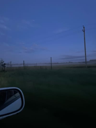 A wide-angle view captures a dimly lit rural landscape at dusk or dawn, likely on the outskirts of Малиновый Куст, Russia. The sky is a deep, clear blue, with faint wisps of clouds visible towards the horizon. A single wooden power pole with electrical lines stretches across the frame, casting a subtle shadow. In the middle ground, a vast, flat field is partially obscured by a low-lying fog, giving the scene a serene and somewhat mysterious atmosphere. Several thin posts or fence markers are visible sparsely across the field. In the foreground, the corner of a car's side mirror is visible, reflecting the blue sky and the edge of the car's interior. The overall impression is one of quiet solitude and the beginning or end of a day in a rural setting.
